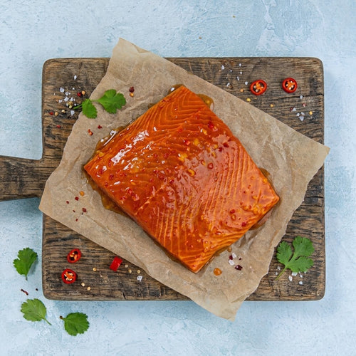 A glazed salmon fillet on parchment paper, placed on a rustic wooden cutting board with scattered herbs, salt, and sliced red chili peppers, set against a light blue textured background.